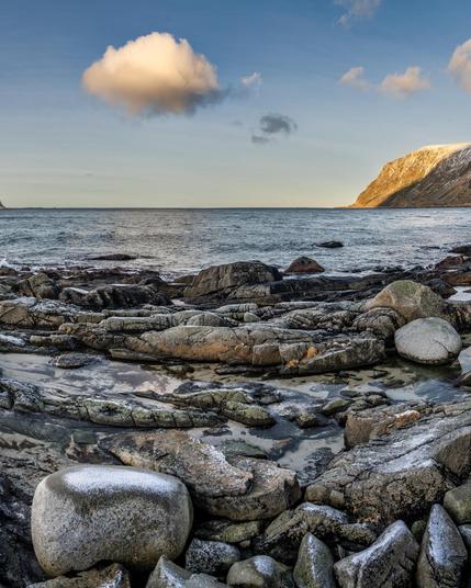 This image depicts a rocky coastal landscape. There are various sizes of rocks in the foreground, some of which are wet, suggesting proximity to the water. The ocean is visible in the middle ground, with calm waves reaching the shore. In the background, a partially snow-covered mountain is seen under a clear blue sky with a few clouds. The lighting suggests it might be early morning or late afternoon.