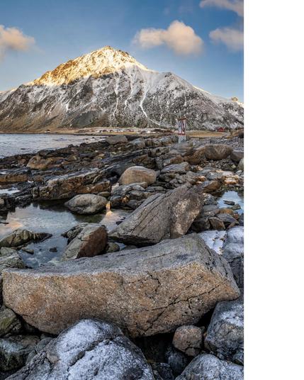 The image features a scenic landscape with a snow-capped mountain in the background. There are large, rugged rocks in the foreground, along with a calm body of water. The sky is clear with a few clouds, creating a serene and picturesque view. A signpost is visible near the center of the image, likely on a coast or shoreline.