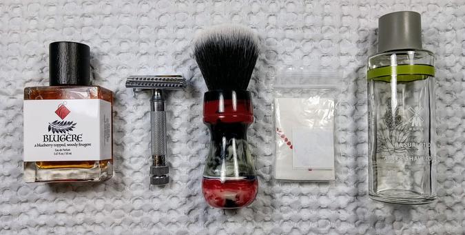 (Le Chat Mistral description)

The image shows five items neatly arranged on a textured white cloth. From left to right: a small bottle labelled "Blugère" with a dark cap, containing a blueberry-topped, woody fragrance. Next, a silver safety razor with a textured handle. Then, a shaving brush with a red handle and black bristles. Beside it, a small packet containing a sample of Mike's Lime shaving soap. Lastly, a clear glass bottle with a metal cap, labelled "Mühle Rasurlotion" for aftershave splash. These items are displayed together but are not necessarily part of a single shaving kit.