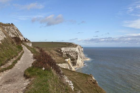 We're on high cliffs, looking along the clifftop with the sea to our right. The path is well worn and set within a narrow boundary of short, pale grass with some dry bushes around. The land rises quickly to the left forming another clifftop set back from the edge ahead and to the left. On the right side the cliff drops to another wide ledge of more grassy land atop a sheer cliff of white chalk. It's then dozens of metres to the sea's surface which appears nearly flat from this distance. It's a bright day with lots of blue sky and little cloud, although some is covering the sun just now, and it feels exposed and windy here.