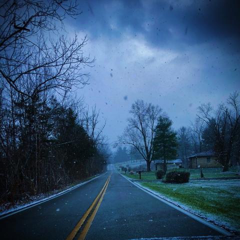 Country road with bare trees on the left and lawns on the right showing green grass peppered with snow. The sky is cloudy, and there is snow on the windshield. Desolation. A long, lonely road home, and the weather shows no signs of improving.