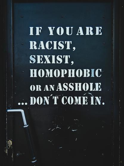 A black door. White letters on it read "If you are racist, sexist, homophobic or an asshole ... don't come in".