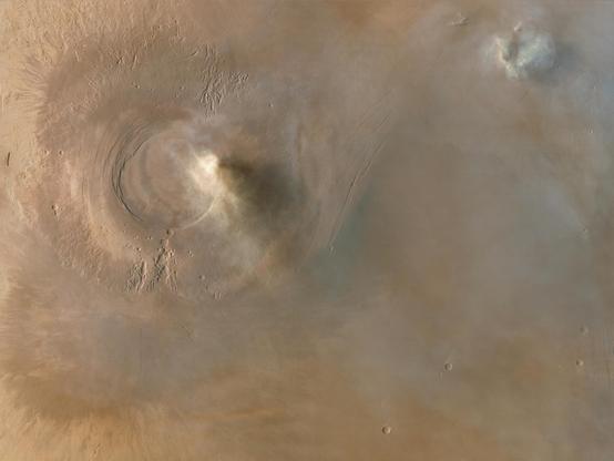 A wider, top-down view of Arsia Mons, a massive Martian volcano, captured by CNSA Tianwen-1 orbiter from an altitude of 688 km on February 7, 2022. The image prominently features the volcano’s summit caldera, surrounded by steep, circular ridges. The rugged, reddish-brown surface is dotted with smaller impact craters and scalloped depressions, likely formed by the collapse of lava tube roofs. Wispy clouds or atmospheric haze partially obscure part of the caldera, casting soft shadows.

In this zoomed-out view, more of Arsia Mons flanks and surrounding terrain are visible. To the upper right, another cloud formation. Faint, dark streaks and long fractures extend across the surface. The location is near 10.3°S latitude and 117.8°W longitude in Mars' western hemisphere. Arsia Mons is the southernmost of the Tharsis volcanoes, spanning 450 km in diameter, rising 20 km high, with a summit caldera 120 km wide