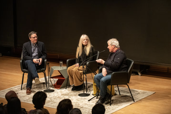 Dan Cavicchi, Tina Weymouth, and Chris Frantz sit together onstage. Frantz is speaking into a microphone and gesturing. The heads of audience members are darkly-lit in the foreground.