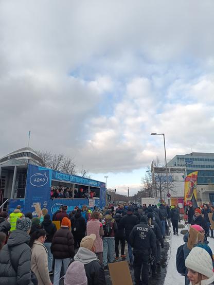 Photo of the winter Pride parade in Berlin. You can see a crowd of people with rainbow flags and the city and sky in the background. There's also a parade truck shown on the left side and the Siegessäule in the distance.