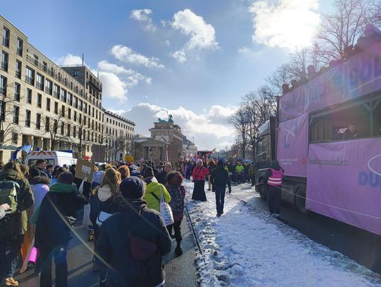 Auf dem CSD: rechts ein Truck mit Musik, links ein Teil der Demo. Im Hintergrund unter blauem Himmel das Brandenburger Tor.