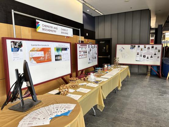 A display area showcasing posters related to chemistry and biochemistry. There are several informational boards, a computer monitor, and models representing molecular structures. Brochures are spread out on a table, along with a promotional banner at the top highlighting the theme.