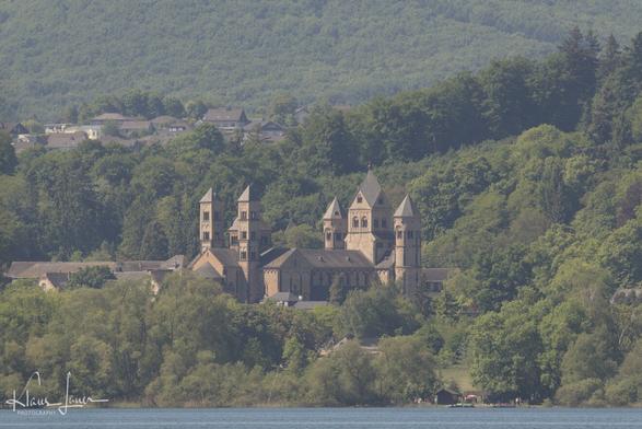 Auf dem Bild sieht man das Laacher Münster, die Abteikirche der Abtei Maria Laach am Laacher See in der Eifel. Das Foto wurde im Frühling mit einem Teleobjektiv von der gegenüberliegenden Seite des Laacher Sees gemacht. Links oberhalb des Klosters sieht man die Ortschaft Bell.