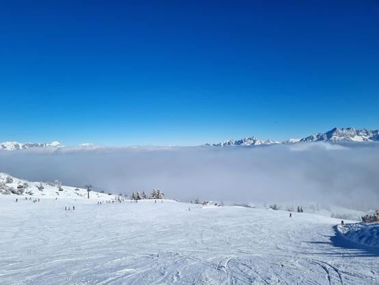 Foto van skipiste met skiërs in de verte, aan de overkant van het dal besneeuwde bergtoppen met daartussen een enorme wolk.
