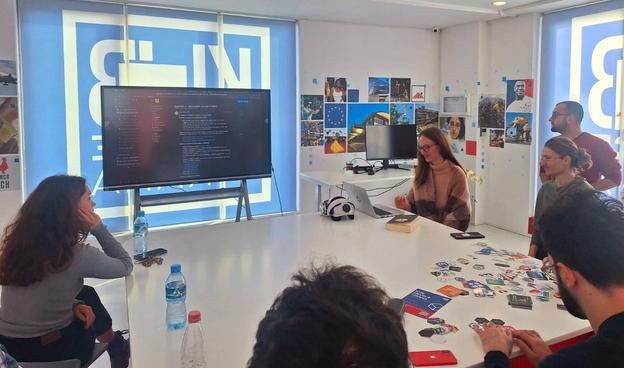 A group of people around a table in front of a large TV. The table has stickers spread. A girl is presenting info that is shown in the TV