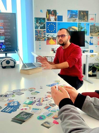 A man is presenting with a laptop in front. On the table stickers are spread.