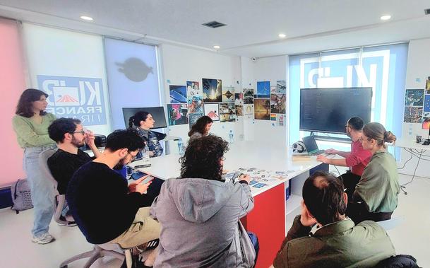 A group of people around a table in front of a large TV. One person on the right is presenting something shown in the large screen.