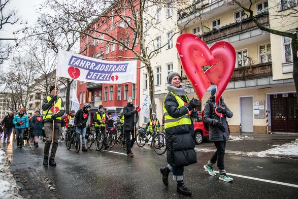 Eine Demonstration läuft durch Schöneberg auf der Straße. Vorn tragen zwei Menschen ein großes, in zwei Stücke zerbrochenes Herz. Dahinter laufen zwei Menschen mit einem großen Banner: "Fahrradstraße jetzt". Sie tragen gebrochene Herzen. Dahinter laufen die Demonstrationsteilnehmenden.