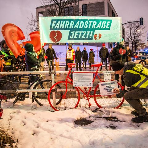 An der Monumentenbrücke steht die Kundgebung. Vorn ist ein knallrotes Rad mit Texten. Auf einem steht, wir warten seit 67 Monaten. Die 67 wird durchgestrichen und durch 68 ersetzt. Dahinter steht die Demonstration mit dem Banner "Fahrradstraße jetzt!" und den großen zerbrochenen Herzen.