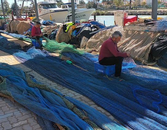 Muelle en un puesto pesquero, al fondo barcos, se ve todo el suelo cubierto de redes grandes alineadas, dos pescadores sentados remiendan las redes.