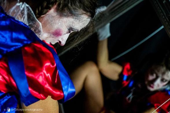 Color photo, landscape format: a female clown, pale and heavily made up, with colorful clothing (Snow White costume), like a marionette, her limbs suspended on strings that protrude upwards out of the picture. She is holding a large mirror with an ornate silver frame in front of her. She is looking into the mirror. The photo was taken from behind the clown, so that the reflection (blurred) is looking at the viewer.
