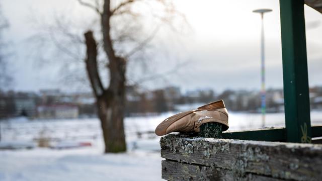 The image shows a pair of beige ballet shoes resting on a wooden ledge. The background features a snow-covered landscape with a tree and a cityscape visible in the distance. The scene appears to be outdoors in winter.