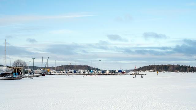 The image shows a wide, snowy landscape with a partially frozen body of water in the foreground. There are several people in the distance, possibly walking or ice skating. In the background, there are trees, buildings, and industrial equipment, like cranes, indicating a harbor or port area. The sky is mostly clear with some clouds.
