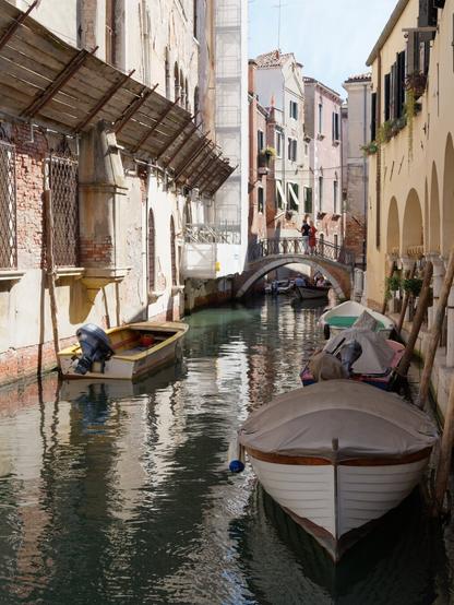The image captures a picturesque canal scene in Venice, Italy. Small boats, some covered and others open, gently float on the calm water, reflecting the historic buildings that line the canal. On the left, an old brick building with arched windows and scaffolding contrasts with the elegant arches and flower-adorned windows of the building on the right. In the background, a charming bridge arches over the canal, where a couple stands, adding a human element to the serene setting. The sunlight casts soft shadows, enhancing the warm and timeless atmosphere of this Venetian alleyway.