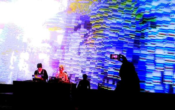 Two performers in front of a large screen with people taking photos at a concert