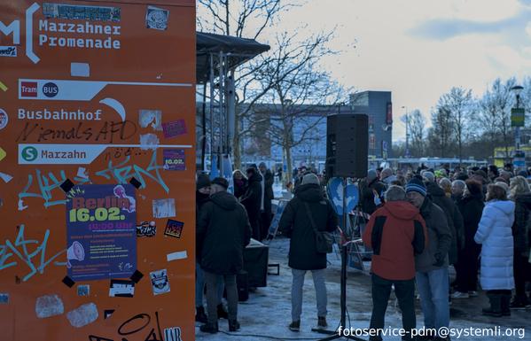auf einer werbetafel, die voll ist mir stickern steht mit einem edding geschrieben "niemals AfD", rechts im bild die AfD Kundgebung