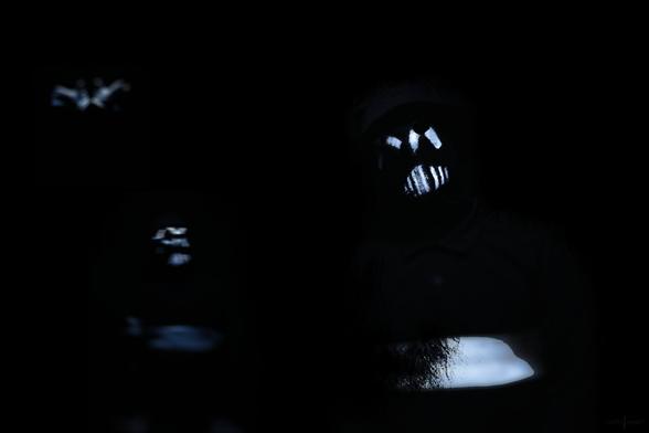 Dark photo shot of the 1 inch army memebers, wearing black clothing with white element stripes and details, also wearing white stripe painted masks, in front of a black background.