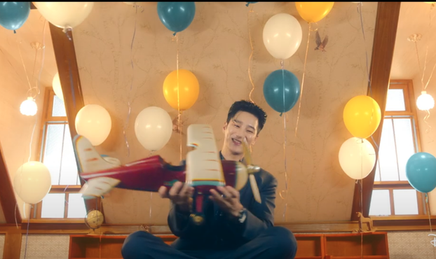 An Bo Hyun in the opening credits for the 2024 South Korean drama "Flex X Cop." He's sitting in a room with blue & yellow balloons around. He's in a blue suit flying a large red toy plane around.