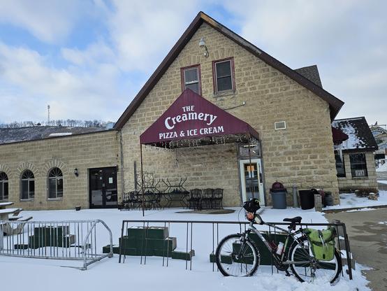 A bicycle with a green bags leaning against a "wheelbender" bicycle parking rack. The ground is white snow. Behind is a brick building with a peaked roof and 4 windows and a double door. On this building is a sign that says "The Creamery Pizza & ice cream.