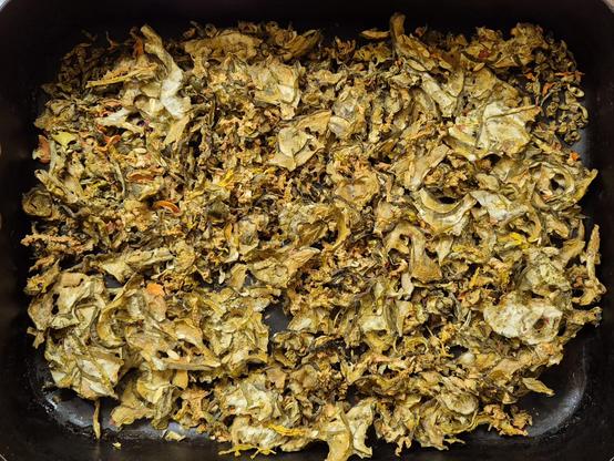 A black roasting tray, filled with olive green flakes of dessicated vegetable matter.