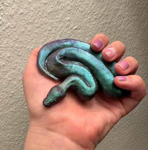Photo of a hand holding a small resin figure of a ball python that is a metallic, iridescent greenish purple.