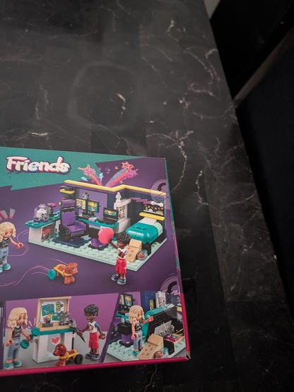 A box with a LEGO Friends set on a dark marble surface features vibrant and playful illustrations. The room setup is primarily in shades of teal, purple, and black, with a bed that has a teal blanket and a desk area that includes two purple chairs with black and teal accents. The computer screens on the desk display colorful graphics. 
, 
, There are two mini-figures depicted. One has light blonde hair, wearing a blue top and light blue pants, and is shown interacting with a small brown dog on wheels. The other mini-figure has dark curly hair, wearing a white shirt with a colorful design and red shorts, and is holding a pink balloon. 
, 
, The background of the box art is a deep purple with colorful star and rainbow graphics, adding to the playful theme. The scenes show the characters engaging in various activities, like working at the desk, playing with the dog, and interacting with the room's accessories. The top left corner of the box has the "Friends" logo in a playful font.