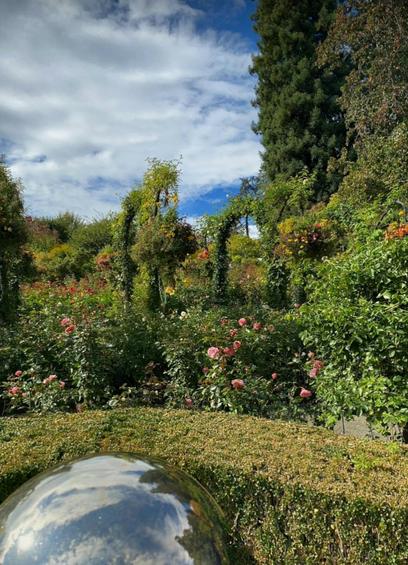 A lush garden with blooming roses, neatly trimmed hedges & archways covered in greenery. A reflective silver sphere sits in the foreground & tall trees stand against a backdrop of a partly cloudy blue sky.