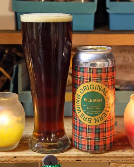 A can of beer with a circular green and yellow label reading “Original Pattern Brewing Wee Man Scotch Ale” stands next to a pilsner glass containing a deep amber fluid with a half-centimeter frothy cream-colored head. The can has a red, yellow, and green tartan pattern.
