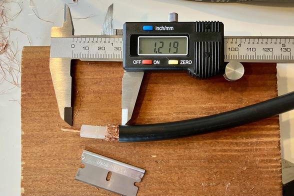 A caliper open to 1.219 inches and the end of a coax cable stripped and ready for crimping, and a razor blade, on a block of wood.