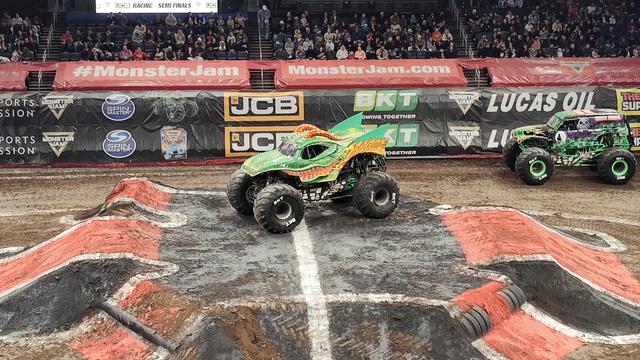 The Dragon monster truck taking a dominant stance in the middle of the arena, on top of the center stunt platform, while Grave Digger lurks in the background