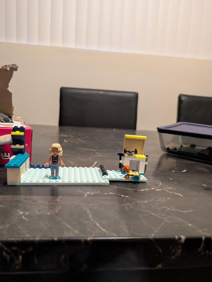 A marble table with a LEGO Friends setup in progress, depicting Nova's Room. On the left side, there is a mini fridge without a door, the beginning of a desk, and a shelf, all in white with blue accents. In front of this section stands the Nova mini doll, who has long blonde hair, a black top with a design, light blue pants, and blue shoes. On the far right is a workbench with a yellow roof and a white base, equipped with tools including a black hammer and a silver wrench, intended for Pickle the dog. Pickle, the orange dog with a blue accessory, is positioned in front of the workbench. The baseplate is light blue, tying the elements together. In the background, there is a pink LEGO box on the left and a clear plastic container with a dark lid on the right. The room has a beige wall with vertical blinds and two black chairs, complementing the overall color scheme.