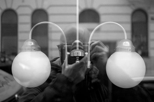 The black and white image shows a close-up of two round light bulbs with frosted glass, mounted on a fixture. The reflection of the photographer, holding a camera, is visible in the glass of the bulbs. The background features a building with arched windows, blurred due to the reflection.