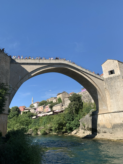 Mostar, an arched stone bridge in BosniaHerzegovina. Tourists on top of the bridge. The river Neretva flows under the bridge. Beautiful Blue skies above. Sunny day. There are dwellings on the sides of the river.