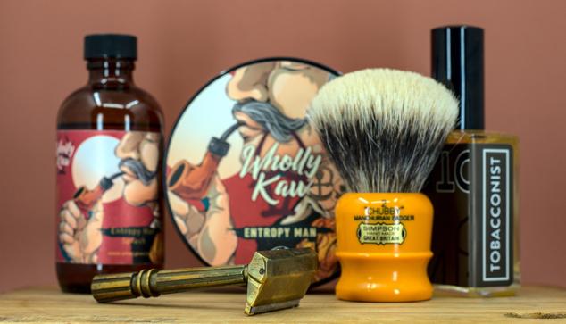 (Le Chat Mistral description)
he image displays five grooming items on a wooden surface against a brown background. From left to right: a brown bottle with a colourful label featuring a muscular figure, labelled "Wholly Kaw Entropy Man." Next, a gold safety razor with an open blade compartment. In the centre, a round shaving soap tin with a matching label. Beside it, a yellow-handled shaving brush with white bristles, labelled "Simpson." Lastly, a small bottle labelled "Tobacconist," likely containing aftershave or fragrance. The items are arranged neatly, suggesting a shaving or grooming setup.