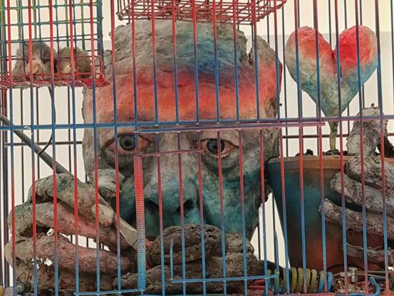 Photo d'une œuvre de Tetsumi Kudo. Une cage métallique colorée en bleu et rouge, renfermant une figure humaine en décomposition aux teintes verdâtres et orangées. Le visage aux yeux expressifs semble émerger de l’intérieur, encadré par des mains squelettiques et terreuses qui manipulent des éléments symboliques. L’une des mains tient un pot contenant une plante en forme de cœur, également colorée de rouge et de bleu. Dans la partie supérieure de la cage, une petite structure grillagée abrite deux formes ressemblant à des créatures hybrides.