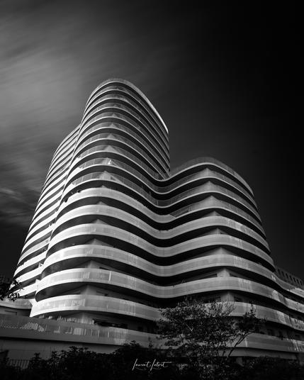 🇫🇷  Un immeuble d'appartements moderne et incurvé avec des balcons superposés, capturé dans un noir et blanc saisissant sur fond de ciel dramatique.

🇬🇧 A modern, curved apartment building with layered balconies, captured in striking black and white against a dramatic sky.
