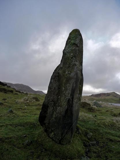 A tall and tapering grey-green standing stone, leaning slightly to the right. It resembles a cowled figure. The stone stands in a rugged landscape of rocky hills, with a lake beyond. The day is overcast and cloudy.