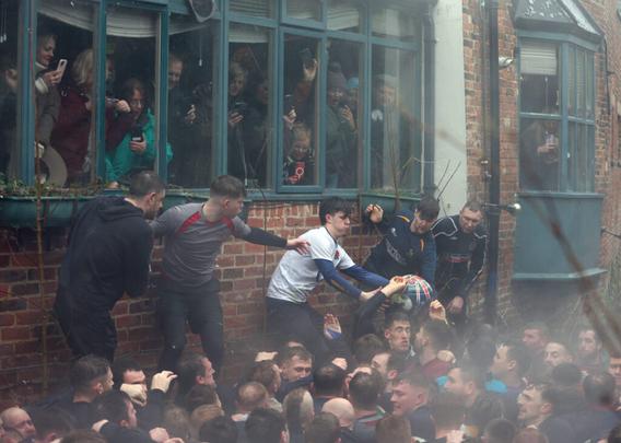 Vor dem Haus kämpfen männlich gelesene Personen um den Shrovetide-Fußball und stehen teilweise auf Pflanzenkübeln. Im Haus schauen vor allem weiblich gelesene Personen am Fenster zu,. Viele haben gerade ihre Handys rausgeholt, um zu filmen. Sie finden es total spannend.