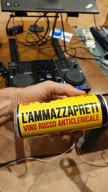 Una lattina con su scritto "l'ammazzapreti, vino rosso anticlericale" e tenuta in mano in primo piano, sullo sfondo una console da Dj.
