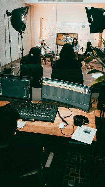 Fotografía. Cabina de grabación de videopodcast, mesa con consola y pantallas en primer plano. Al fondo se encuentra un cuarto insonorizado con cinco personas, tres de ellas frente a las cámaras.