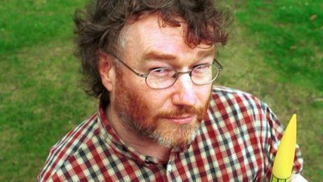 A head and shoulders photo of Iain Banks. He has brown, curly, untidy hair, and a reddish beard beginning to whiten on his chin. He is wearing glasses and a red, white, and blue checked shirt. He's looking up at the camera, which is above head height; we only see grass behind him. He appears to be carrying a toy rocket: its yellow nose-cone pokes up into the bottom of the photograph.