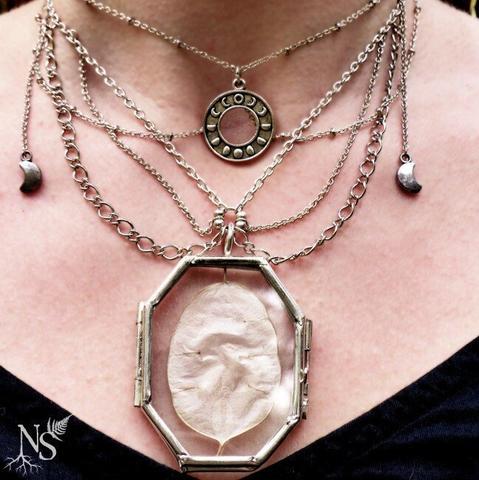 English A close up on an intricate necklace with cascades of silver chains, a round charm with the Moon cycles and 2 tiny Moon charms falling on the sides. The centerpiece is a glass medallion showcasing a lunaria annua disc that looks a lot like a full Moon.
It is worn on a neck and the shirt neckline at the bottom is black.
❉
Français Gros plan sur un collier complexe avec des cascades de chaînes en argent, une breloque ronde avec les cycles de la Lune et 2 petites breloques Lune tombant sur les côtés. La pièce maîtresse est un médaillon en verre présentant un disque de Lunaria annua qui ressemble beaucoup à une pleine Lune.
Il est porté autour du cou et le col du vêtement en-dessous est noir.