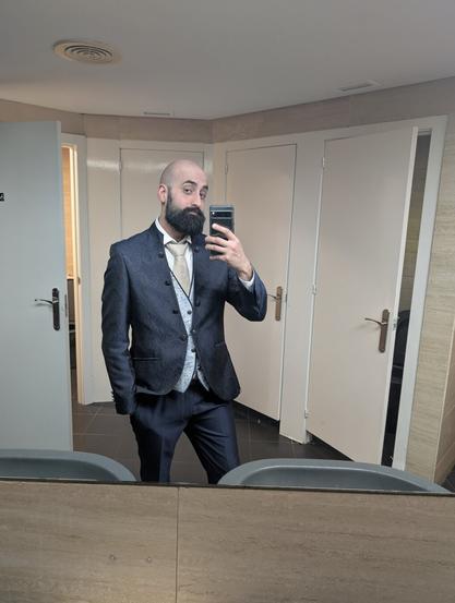 A selfie of a man, he has shaved head, a beard, and is wearing a navy blue jacket with a paisley pattern on it. There's a vest under it, with the same patter but in sky blue colour, a white shirt, and a matte golden/beige tie. It's a very nice man wearing a very nice suit.