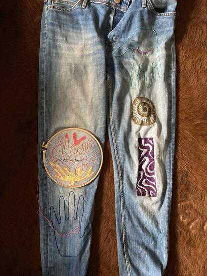 Work in progress for a visible mending on the right knee of a levi's jeans. The embroidery represents a coral with shades of yellow, orange and red threads. The rst of the jeans has more embroideries. On the left leg there is a purple abstract rectangular embroidery.