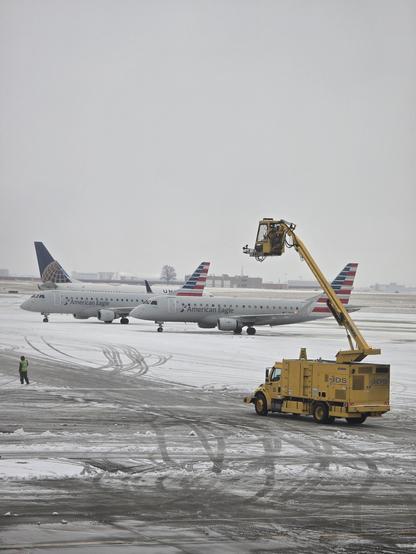 A man is seen working on a lift attached to a plane at an airport. In the background, there are two American Eagle planes parked on the tarmac. One of the planes has a crane nearby. The scene also includes a large machinery vehicle and a truck. The runway appears to be snowy, adding to the wintery setting. The man on the lift is focused on his task, ensuring the plane is maintained properly. Overall, the image captures a busy and cold day at the airport with various vehicles and planes in operation.